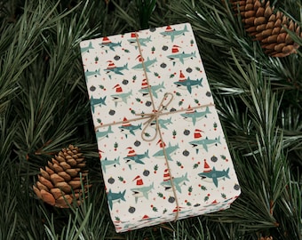 May include: A white gift box wrapped in festive wrapping paper featuring a repeating pattern of blue sharks wearing Santa hats. The paper is tied with twine and the box is sitting on a bed of green pine needles.