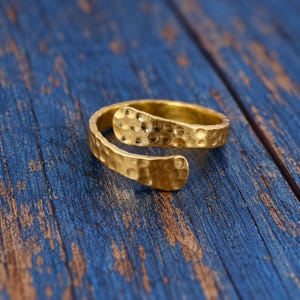 May include: A gold-toned open ring featuring a textured, hammered surface finish. The band is adjustable, with two rounded, overlapping ends that display a consistent dimpled pattern across the entire exterior of the metal jewelry piece.