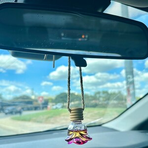 Pode incluir: Um aromatizador de carro de vidro rosa e amarelo em forma de flor pendurado no espelho retrovisor de um carro. O aromatizador est&aacute; preso a uma corda marrom com contas de madeira.