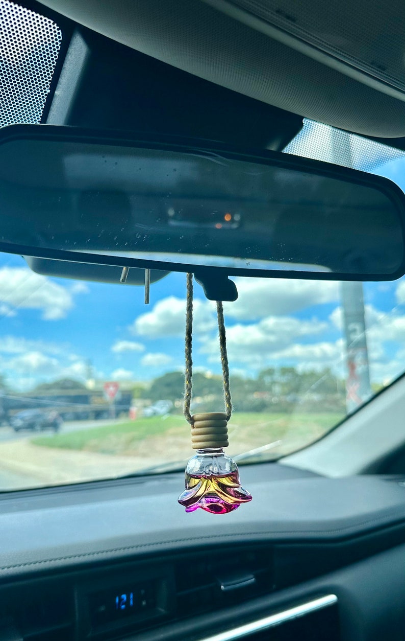 Pode incluir: Um aromatizador de carro de vidro rosa e amarelo em forma de flor pendurado no espelho retrovisor de um carro. O aromatizador est&aacute; preso a uma corda marrom com contas de madeira.