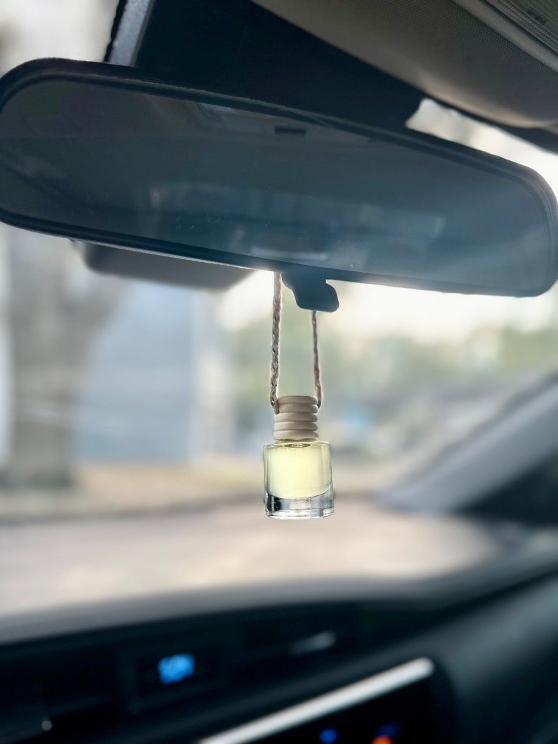 May include: A small glass bottle with a light yellow liquid hangs from a rearview mirror in a car. The bottle has a wooden cap and a string attached to it.