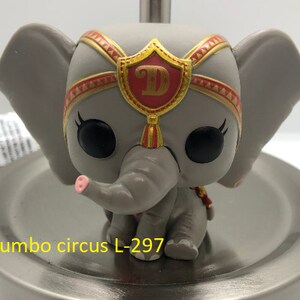 May include: A gray and pink Dumbo circus elephant figurine with a red and gold hat. The hat has a gold shield with a letter D on it. The figurine is sitting on a silver surface.