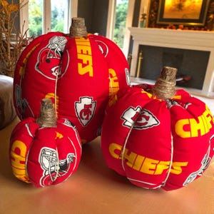 May include: Three fabric pumpkins in varying sizes featuring a red background with yellow text reading CHIEFS and white Kansas City Chiefs football team logos. Each pumpkin has a textured brown stem wrapped in thin twine and vertical white stitching.