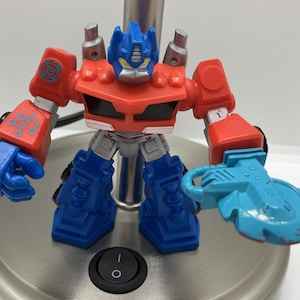 May include: A red and blue Transformers action figure with a blue saw blade in its right hand. The figure has a red chest and blue legs. The figure is standing on a silver surface.