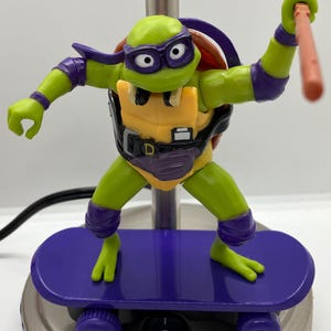 May include: A green Teenage Mutant Ninja Turtles action figure on a purple skateboard. The figure is wearing a purple mask, a yellow and purple shell, and a purple bandana. The figure is holding a red staff.