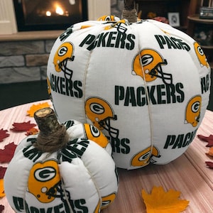 May include: Two fabric pumpkins featuring a white background with a repeating pattern of yellow and green Green Bay Packers football helmets and the word PACKERS in green text. Each pumpkin has a natural twig stem wrapped with brown twine.