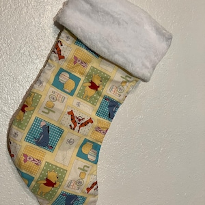 May include: A yellow Christmas stocking with a white faux fur cuff. The stocking features a patchwork design with Winnie the Pooh and his friends, including Tigger, Piglet, and Eeyore. The stocking is filled with a variety of colorful patterns, including polka dots, stripes, and floral designs.
