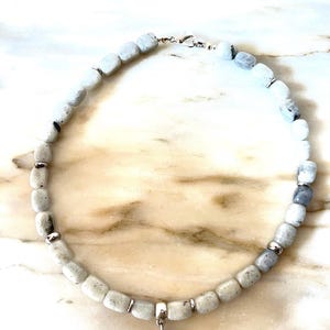 May include: A beaded necklace featuring light gray and white square beads with silver accents. A polished, heart-shaped pendant hangs from the center. The necklace is displayed on a marble surface.