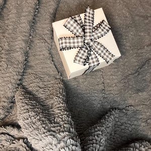 May include: A white gift box with a black and white checkered ribbon tied in a bow. The box is sitting on a gray fuzzy blanket.