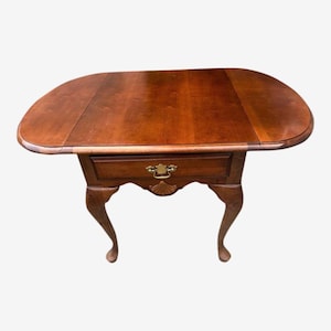 May include: A wooden drop-leaf table with a rich, warm brown finish. The table features a single drawer with a decorative brass handle and cabriole legs. The table measures approximately 36 inches long when open.
