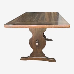 May include: A dark brown wooden table with a rectangular top and a unique pedestal base. The table's top appears to be approximately 60 inches long and 36 inches wide, with a lighter-colored edge. The base has an ornate, carved design.