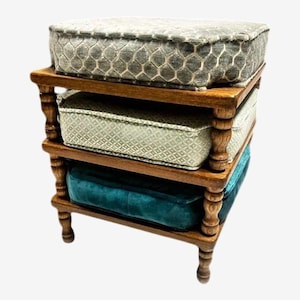 May include: A wooden, three-tiered shelf unit with turned legs. Each shelf holds a plush, square cushion in varying colors: gray, light green, and teal. The cushions have patterned designs, creating a layered, decorative storage solution.