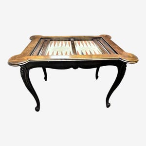 May include: A vintage backgammon table with a dark wood frame and legs. The playing surface features a light-colored field with alternating triangles in shades of brown and white. The table has a classic, elegant design.