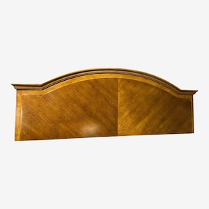 May include: A wooden headboard with a curved top and a warm, golden-brown finish. The headboard features a symmetrical design with a central panel and a decorative border. The wood grain is visible, adding to its classic appeal.