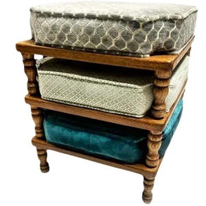 May include: A three-tiered wooden shelf unit with three upholstered cushions. The top cushion is gray with a geometric pattern, the middle is light green, and the bottom is teal. The wooden frame has turned legs and supports.