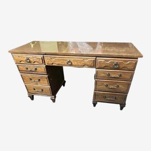 May include: A wooden desk with a light brown finish. It features multiple drawers on both sides, each with ornate handles. The desk has a rectangular top and is supported by four legs.