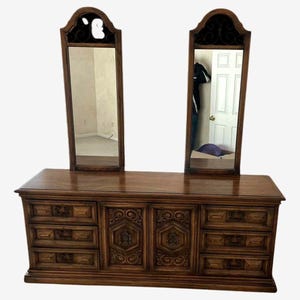 May include: A dark brown wooden dresser with two attached mirrors. The dresser has multiple drawers and ornate carvings on the cabinet doors. The mirrors have decorative tops.