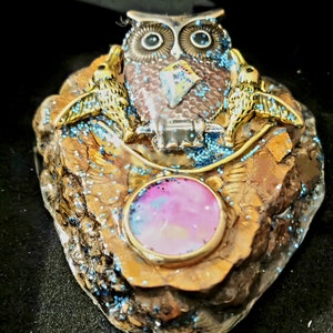 May include: A resin owl figurine with large eyes and a diamond-shaped gem on its chest, perched on a brown and gold rock with blue glitter, and a pink gem in the center.