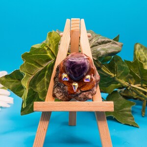 May include: A purple amethyst stone set in a wooden owl-shaped pendant with iridescent rhinestones. The pendant is displayed on a wooden easel against a blue background.