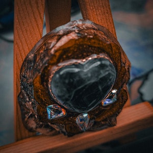 May include: A heart-shaped resin piece with a dark gray center and clear rhinestones around the edges. The resin is set in a brown, textured, wooden frame.