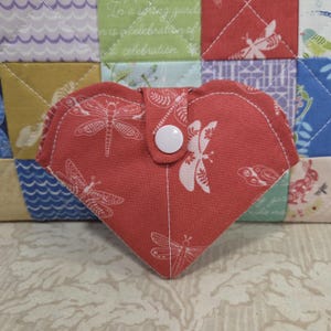 May include: A heart-shaped, coral-colored fabric item with a white dragonfly and butterfly pattern. It has a white button closure and is likely a small pouch or case. The background features a patchwork quilt.