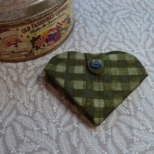 May include: A heart-shaped fabric pouch featuring a dark and light green plaid pattern, secured with a single dark button at the top center. The pouch is positioned near a round metal tin with a Santa Claus illustration and text.