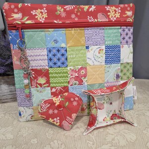May include: A quilted patchwork zippered pouch with a red floral top and zipper, accompanied by a heart-shaped item and a small tray. A beaded keychain hangs from the zipper. The items feature a variety of colorful floral and geometric patterns.