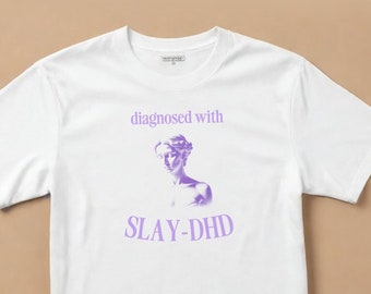 May include: White t-shirt with a crew neck featuring the text "diagnosed with SLAY-DHD" in purple, along with a purple illustration of a classical bust. The shirt is made of a soft, comfortable material.