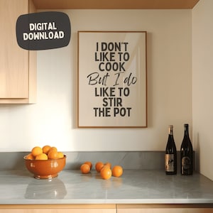 Pode incluir: Uma impressão emoldurada com o texto "Não gosto de cozinhar, mas gosto de mexer na panela" pendurada em uma parede de uma cozinha. Há laranjas em uma tigela e duas garrafas de vinho no balcão.