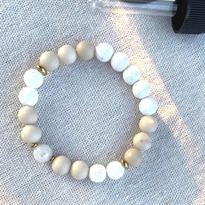 May include: A bracelet made of alternating white and beige round beads, with gold-coloured spacers. A dropper bottle is visible in the background. The beads are likely 0.8 cm in diameter.