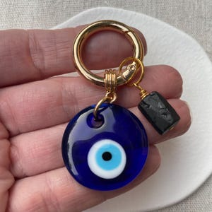 May include: A blue glass Evil Eye charm with a white and blue iris, attached to a gold-colored keyring. A small black rectangular bead hangs from the charm. The charm is designed to ward off negative energy.
