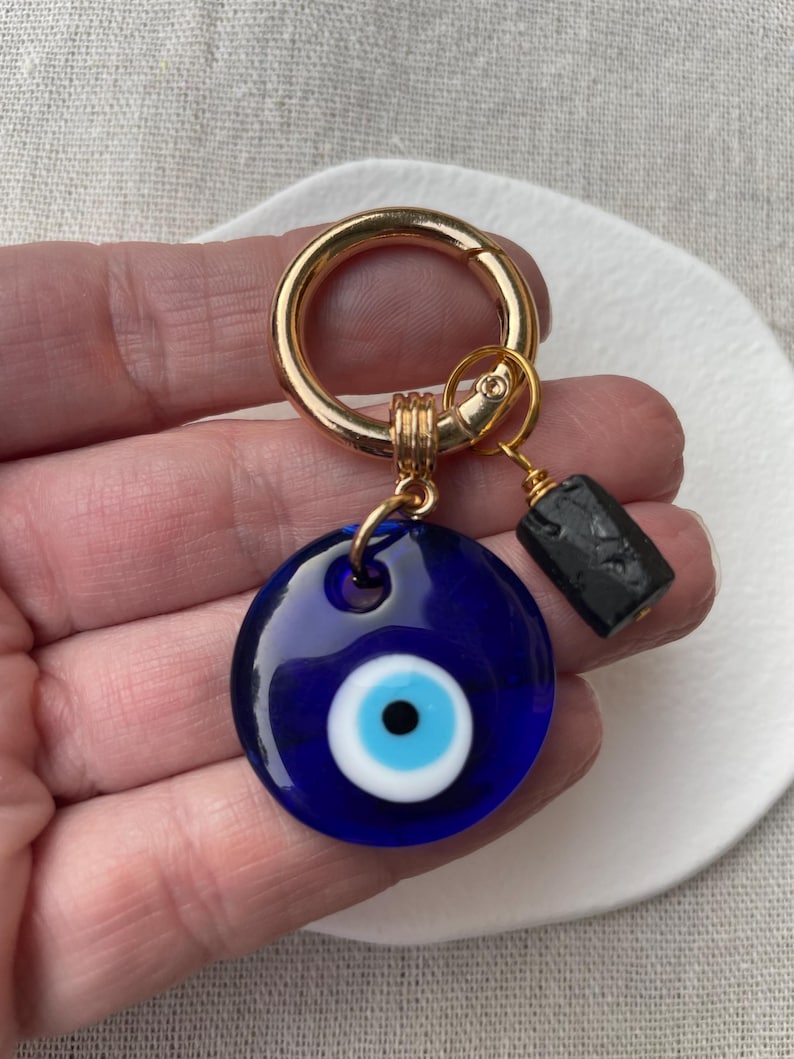 May include: A blue glass Evil Eye charm with a white and blue iris, attached to a gold-colored keyring. A small black rectangular bead hangs from the charm. The charm is designed to ward off negative energy.