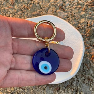 May include: A round, dark blue keychain charm with a gold-colored clasp. The charm features a white and light blue eye design. The keychain is a protective amulet, often used for good luck and warding off negative energy.