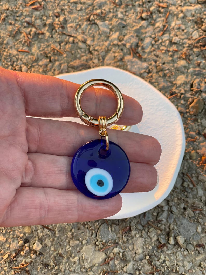 May include: A round, dark blue keychain charm with a gold-colored clasp. The charm features a white and light blue eye design. The keychain is a protective amulet, often used for good luck and warding off negative energy.