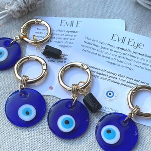 May include: Close-up of several blue Evil Eye keychains with gold-colored hardware. Each keychain features a round, dark blue charm with a white and light blue eye design. Small black stone accents are also present.