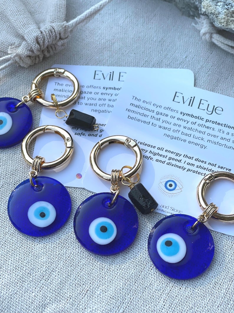 May include: Close-up of several blue Evil Eye keychains with gold-colored hardware. Each keychain features a round, dark blue charm with a white and light blue eye design. Small black stone accents are also present.