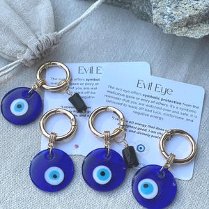 May include: Four blue Evil Eye keychains with gold-colored rings and black accents. Each keychain features a circular blue charm with a white and blue eye design. Cards with text about the Evil Eye are also visible.