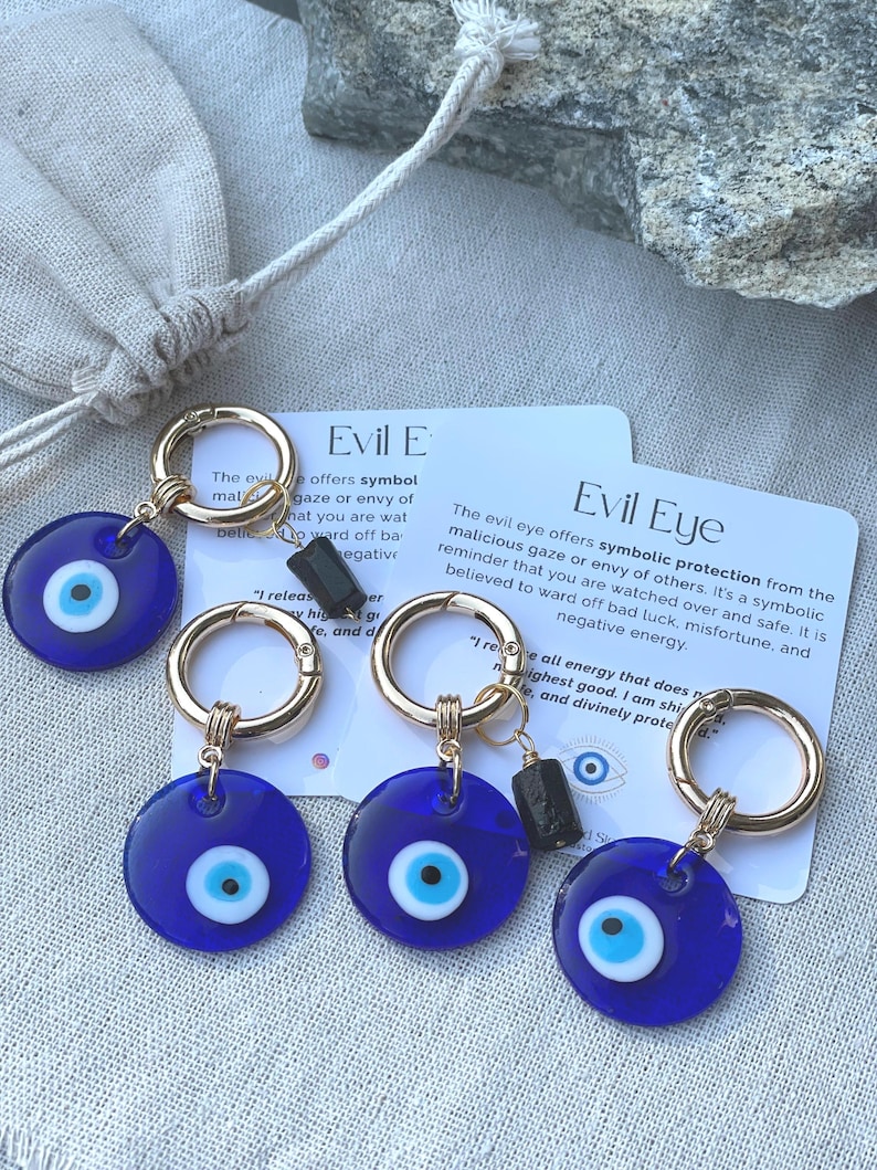 May include: Four blue Evil Eye keychains with gold-colored rings and black accents. Each keychain features a circular blue charm with a white and blue eye design. Cards with text about the Evil Eye are also visible.