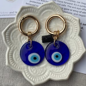 May include: Two blue evil eye keychains with gold-colored clasps. Each keychain features a round, dark blue charm with a light blue and white eye design. The keychains are displayed on a white, decorative plate.