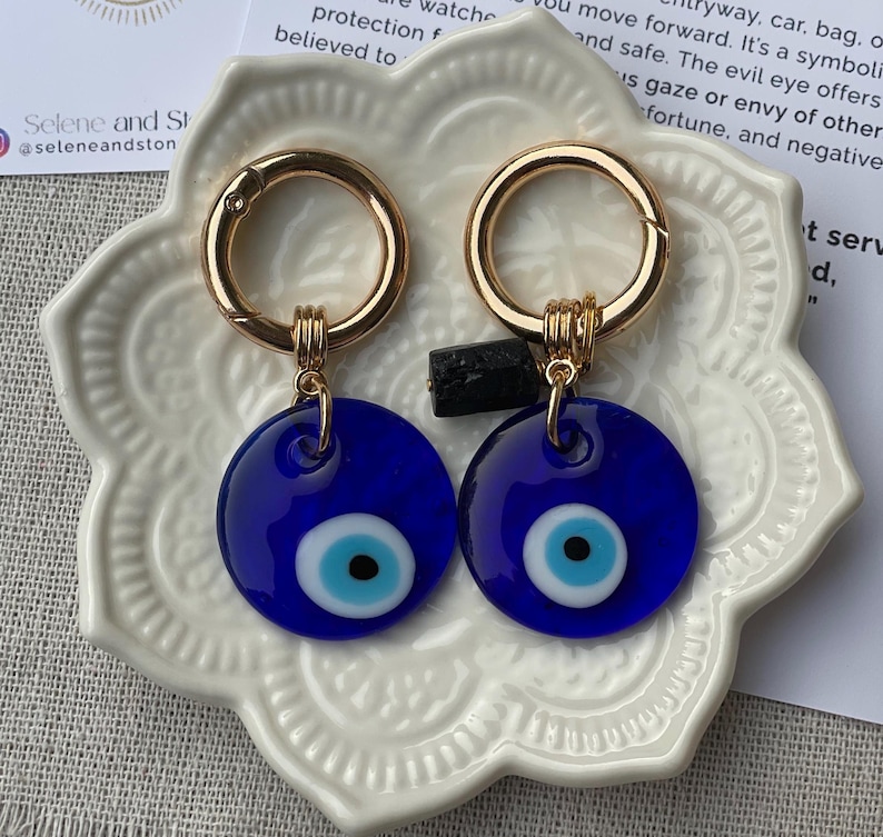 May include: Two blue evil eye keychains with gold-colored clasps. Each keychain features a round, dark blue charm with a light blue and white eye design. The keychains are displayed on a white, decorative plate.