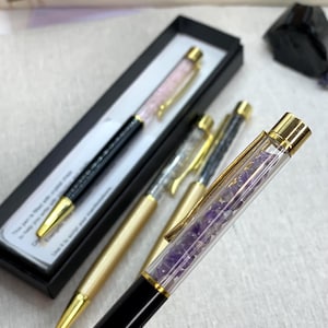 Peut inclure: Stylos élégants avec des accents de cristal. Un stylo a un corps noir avec des garnitures dorées et un corps rempli de cristaux roses. Un autre stylo a un corps doré avec un corps rempli de cristaux transparents. Un troisième stylo a un corps noir avec un corps rempli de cristaux violets.