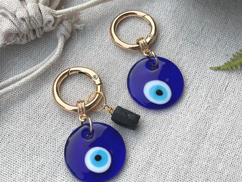 May include: Two gold-colored keychains with blue evil eye charms. Each charm has a white and light blue eye design. One keychain has a small black stone attached. The keychains are on a textured, light-colored surface.