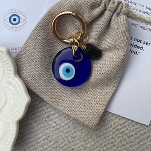 May include: A blue evil eye charm keychain with a gold-colored ring and a small black stone. The charm is round with a white and light blue eye design. The keychain is on a beige drawstring bag.