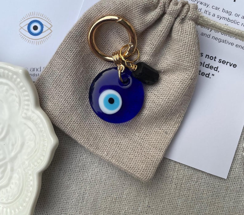 May include: A blue evil eye charm keychain with a gold-colored ring and a small black stone. The charm is round with a white and light blue eye design. The keychain is on a beige drawstring bag.