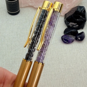 May include: Two pens with gold-coloured barrels and clips. One pen is filled with black stones, the other with purple stones. The pens are displayed with a bottle of essential oil, a rose quartz crystal, and other gemstones.