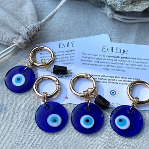 May include: Several blue Evil Eye keychains with gold-colored clasps and black accents. Each keychain features a round, dark blue charm with a white and light blue eye design. The keychains are displayed on a neutral-toned surface.