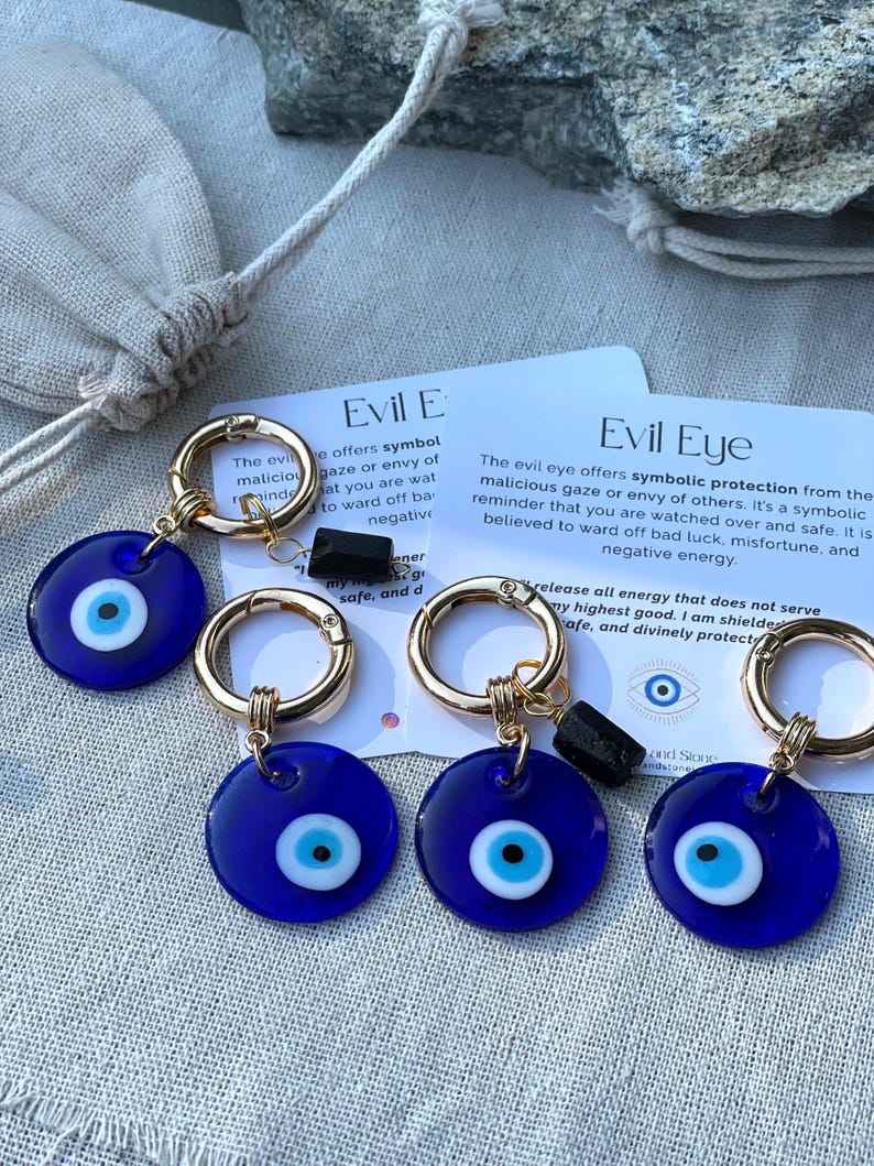May include: Several blue Evil Eye keychains with gold-colored clasps and black accents. Each keychain features a round, dark blue charm with a white and light blue eye design. The keychains are displayed on a neutral-toned surface.