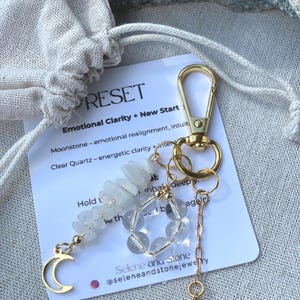 Puede incluir: Un llavero dorado con un diseño de cuentas de cuarzo transparente y piedra lunar, un dije de media luna dorada y un dije con la letra "M" dorada. El llavero está sujeto a una tarjeta con la palabra "RESET" y texto.