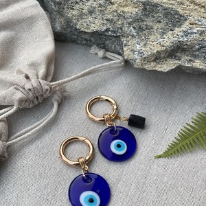 May include: Two gold-colored hoop earrings with a dark blue, circular charm featuring a white and black eye design. A small, black rectangular stone hangs from one earring. The earrings are displayed on a neutral-colored surface.