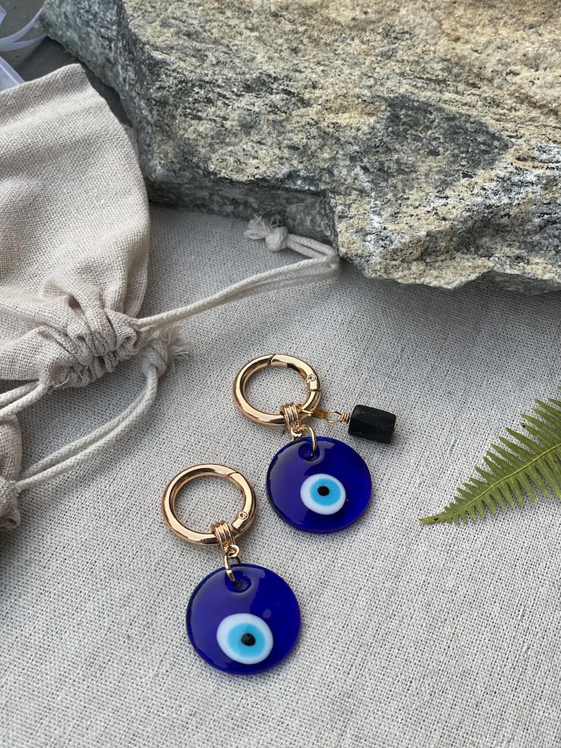 May include: Two gold-colored hoop earrings with a dark blue, circular charm featuring a white and black eye design. A small, black rectangular stone hangs from one earring. The earrings are displayed on a neutral-colored surface.